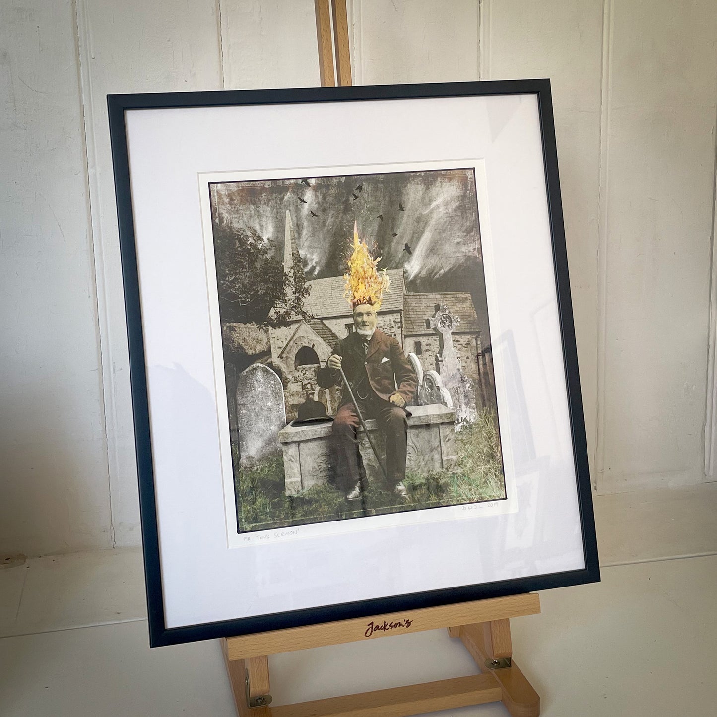 Framed artwork of a person with a hat on an easel against a white wall.