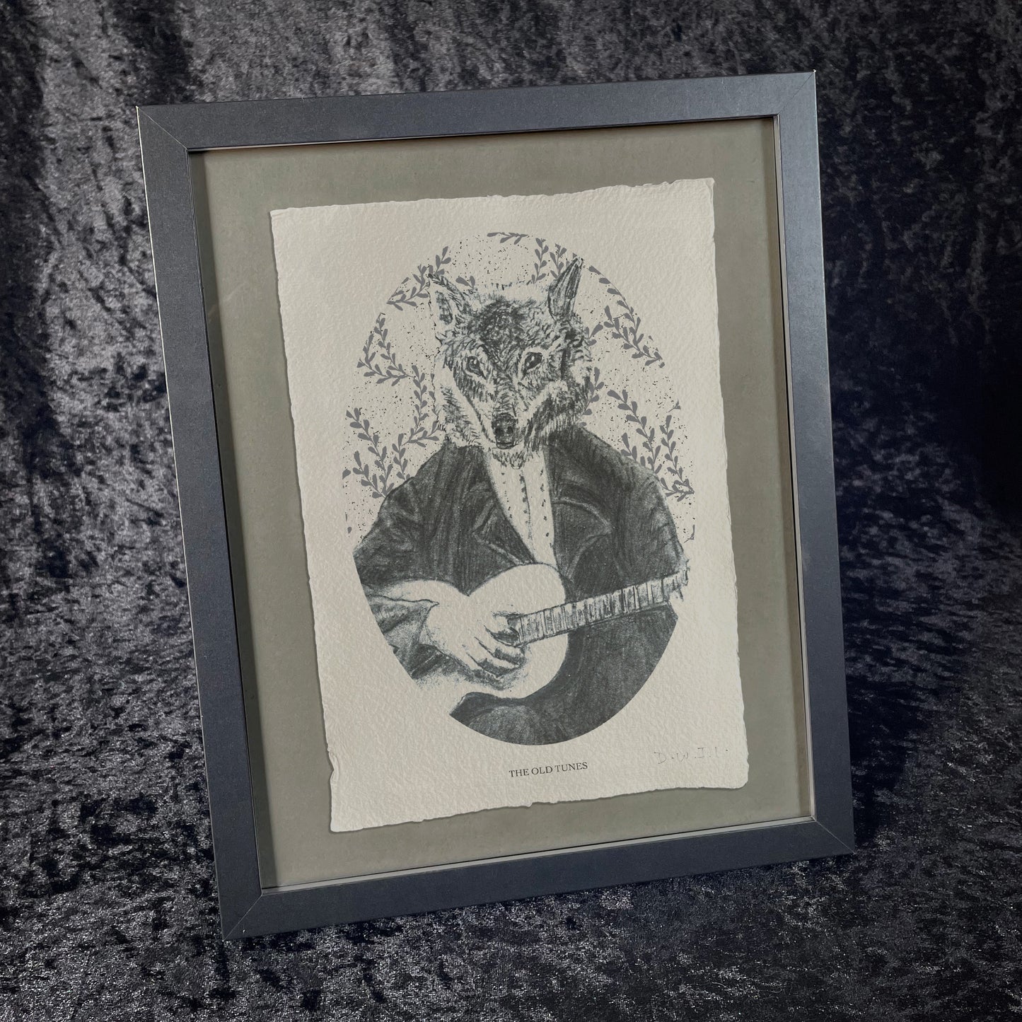 Framed black-and-white printed illustration of a wolf dressed in Victorian attire, playing a stringed instrument. The background features leafy patterns, and the print sits on textured paper against a dark, velvety fabric backdrop.