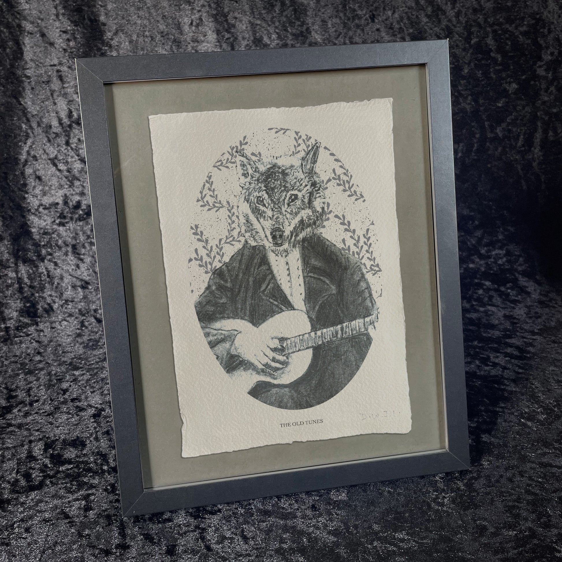 Framed black-and-white printed illustration of a wolf dressed in Victorian attire, playing a stringed instrument. The background features leafy patterns, and the print sits on textured paper against a dark, velvety fabric backdrop.