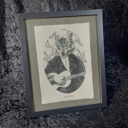 Framed black-and-white printed illustration of a wolf dressed in Victorian attire, playing a stringed instrument. The background features leafy patterns, and the print sits on textured paper against a dark, velvety fabric backdrop.